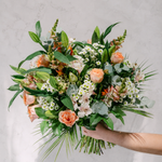 A vibrant floral bouquet held by a person's hand against a light gray marble background. The bouquet features peach roses, white daisies, green foliage, and eucalyptus leaves.