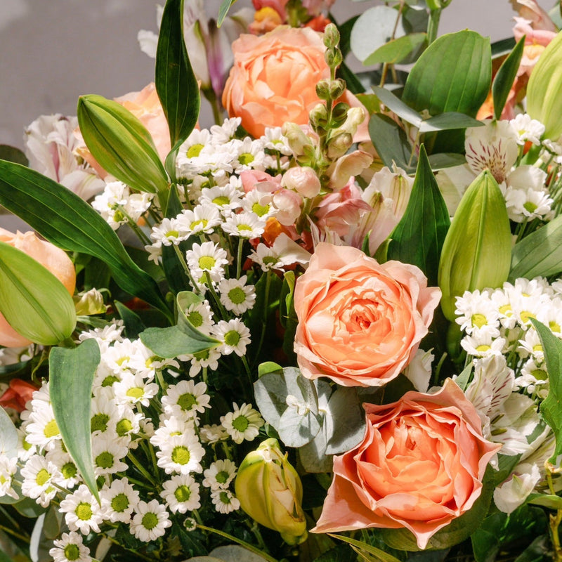 A close-up of a floral arrangement featuring peach roses, white daisies, green foliage, and soft pink flowers in a glass container.