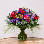 A vibrant artificial flower bouquet in a clear glass vase, featuring red and pink roses, purple and blue filler flowers, green eucalyptus, and palm leaves, displayed on a wooden table against a light textured wall.
