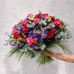 A vibrant, large bouquet of fresh flowers held by a person's arm against a light gray marble background. The bouquet features a rich mix of pink and red roses, deep purple and blue filler flowers, green eucalyptus leaves, and long green palm fronds.