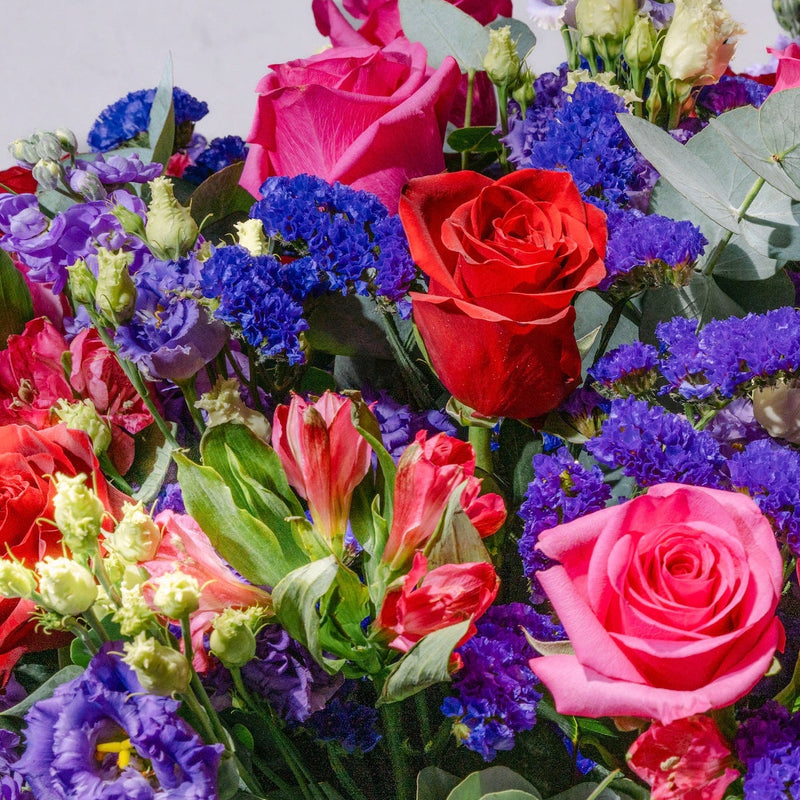 A vibrant mixed flower bouquet featuring pink and red roses, blue and purple flowers, green foliage, and red alstroemeria, set against a plain light gray background.