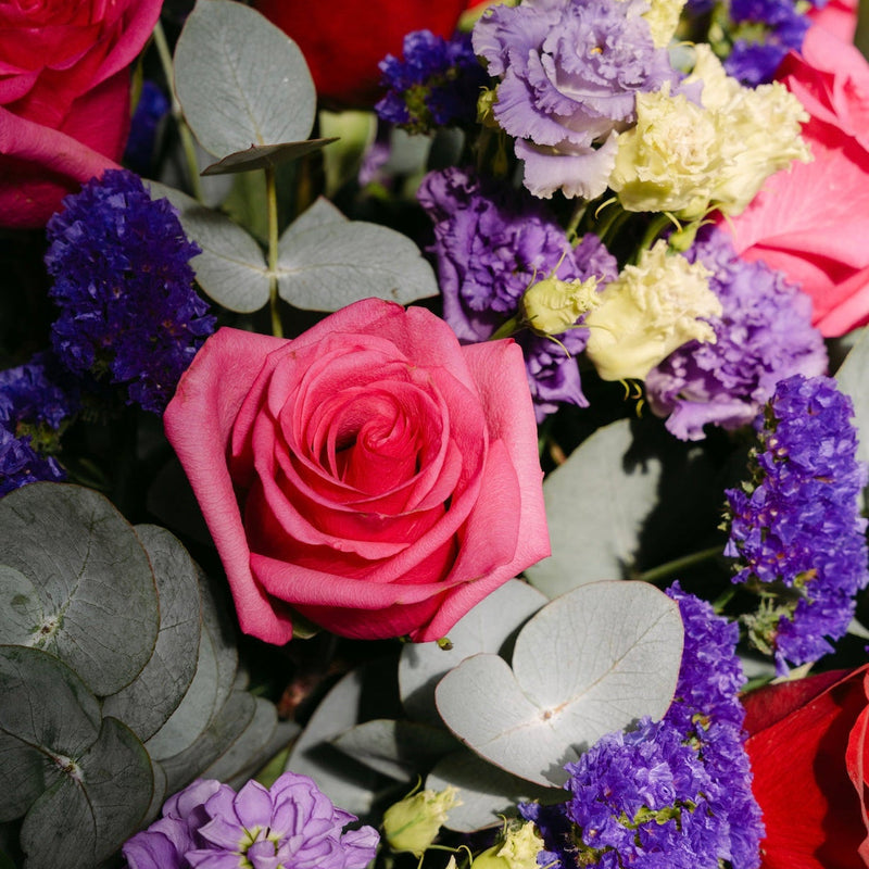 A close-up of a vibrant floral arrangement featuring pink and red roses, purple and lavender hydrangeas, eucalyptus leaves, and small pale yellow flowers.